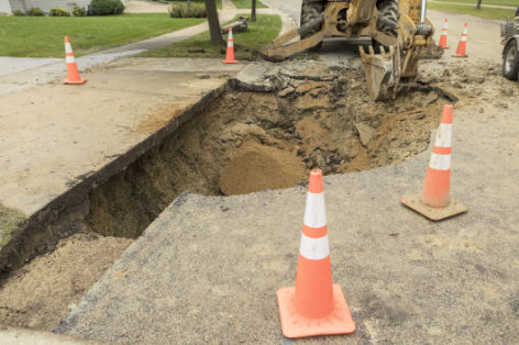 A large hole in a residential street, near a house driveway, the result of a broken water main. A backhoe is being used to remove damaged asphalt.